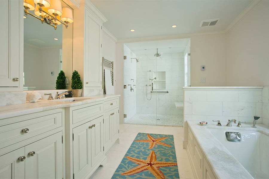 Coastal bathroom with off-white cabinetry, marble countertops and backsplash, glass door shower, and decorative rug