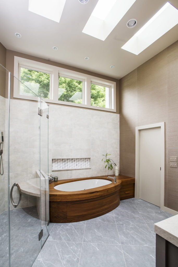 Modern bathroom with wooden bathtub, all glass shower, large grey tile flooring, beige paneling on walls, and marble tile backsplash