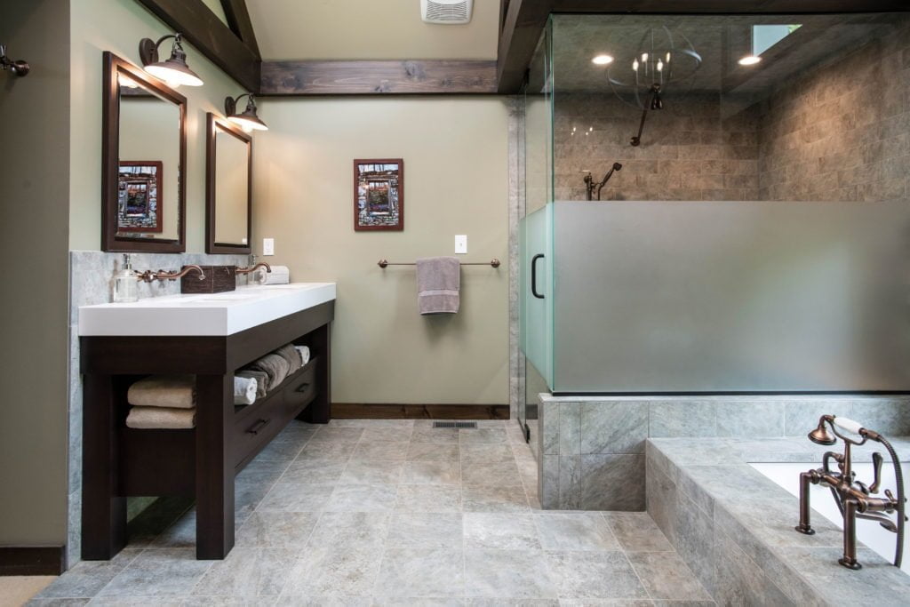 Rustic bathroom with grey tile flooring, dar hardwood cabinetry and trim, dark beige wall color, all glass shower, and marble countertops