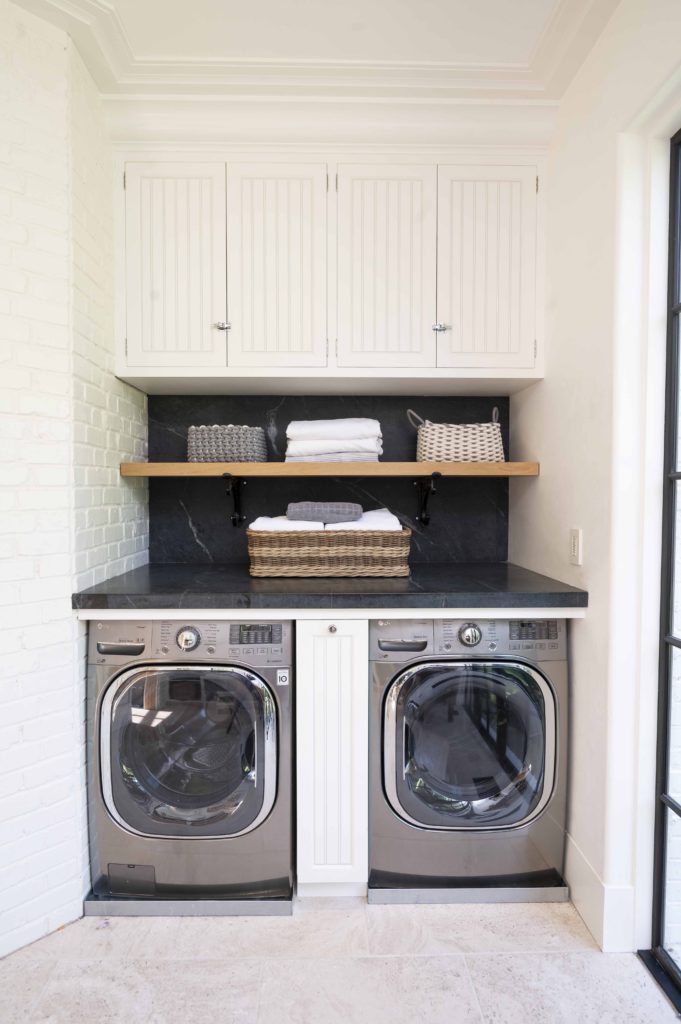 Coastal laundry room with white paneled cabinetry, dark granite countertops, and wooden shelving