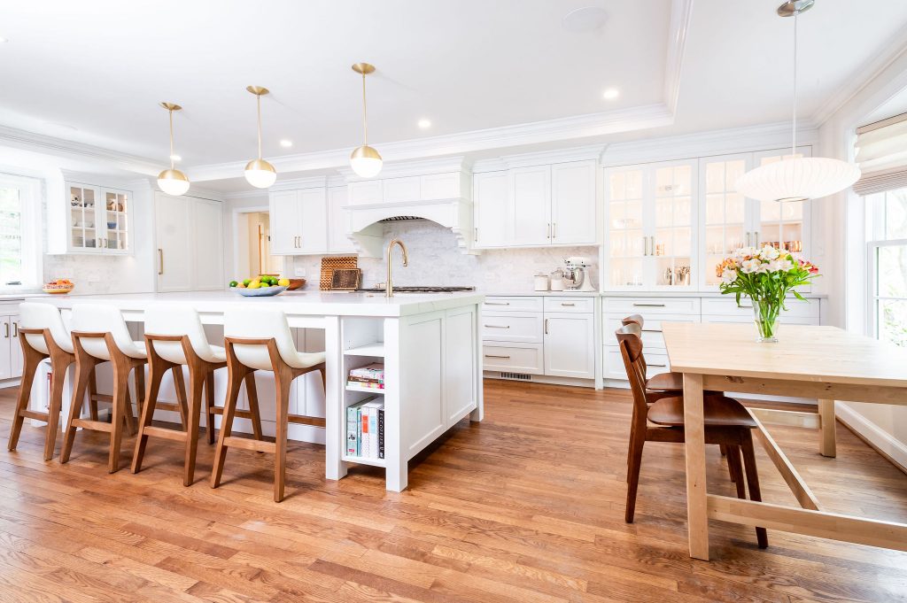 Transitional kitchen/dining room with white cabinetry, marble countertops, medium hardwood flooring, wooden hi-top and dining chairs, and a wooden dining table, gold accent